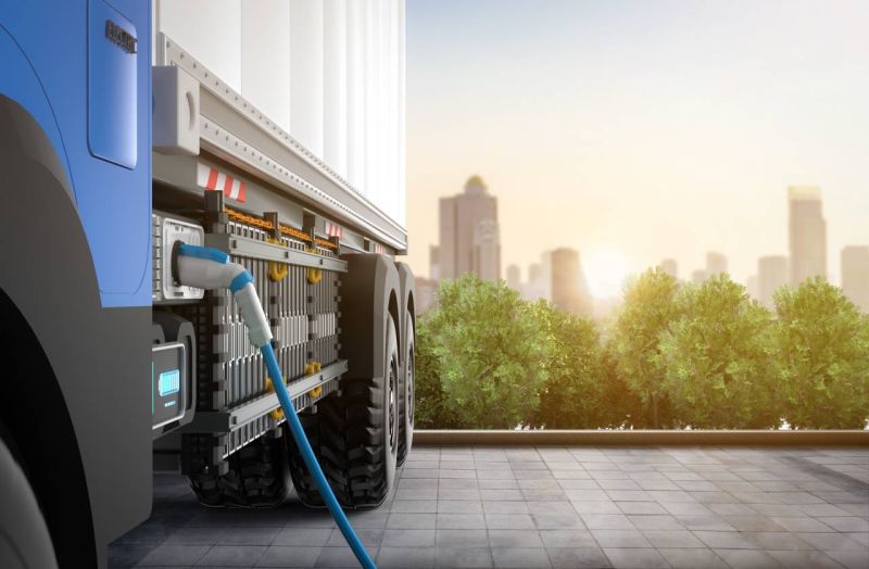 An electric lorry fitted with a charging hose, parked in front of a landscape with trees in the foreground and tower blocks behind.